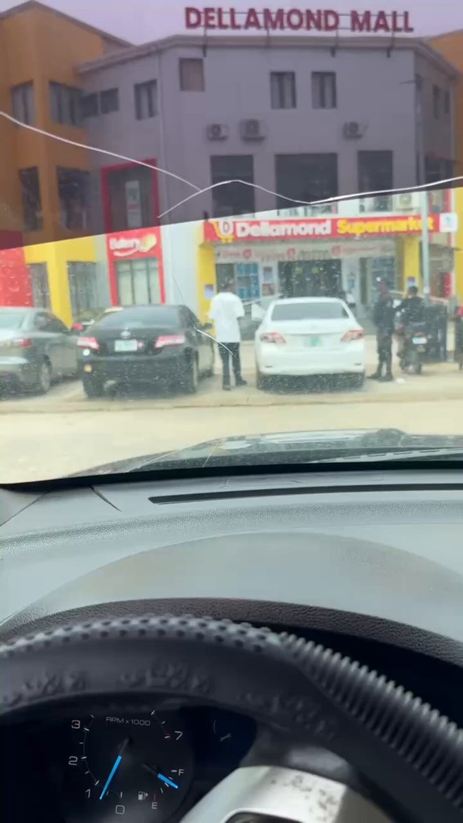 The image shows a car's windshield with a clock displaying the time as 12:30 pm. The scene outside the car window features several people standing near a building, possibly a shopping center or a market. There are also multiple cars parked nearby, including one that appears to be a black sedan and another white car.