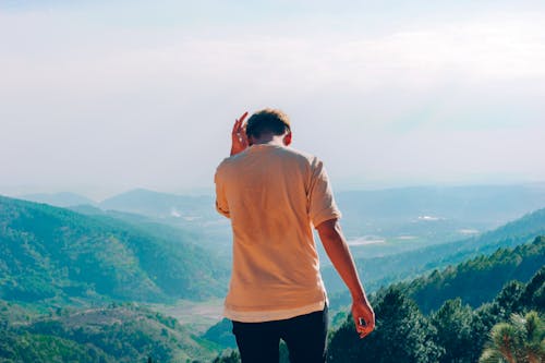 The image shows a man standing at the top of a mountain, overlooking a valley below him. The sky above them appears hazy, suggesting that they are in a mountainous area with potentially poor air quality due to pollution or smog. The man is wearing a white shirt and black pants while looking out over the landscape.