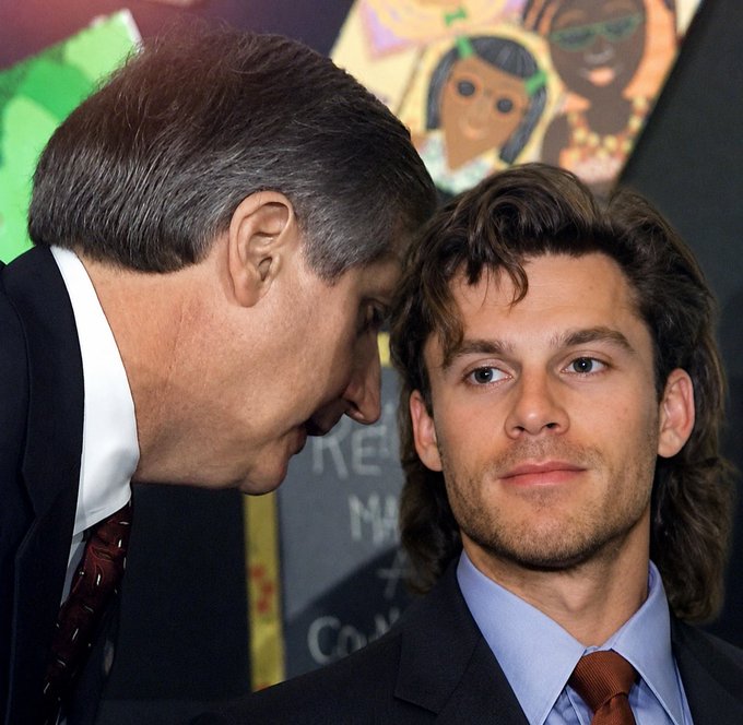 Two men in suits are pictured at what appears to be a public event, with an older man leaning in to whisper to a younger man with longer brown hair; part of a colorful poster is visible in the background.