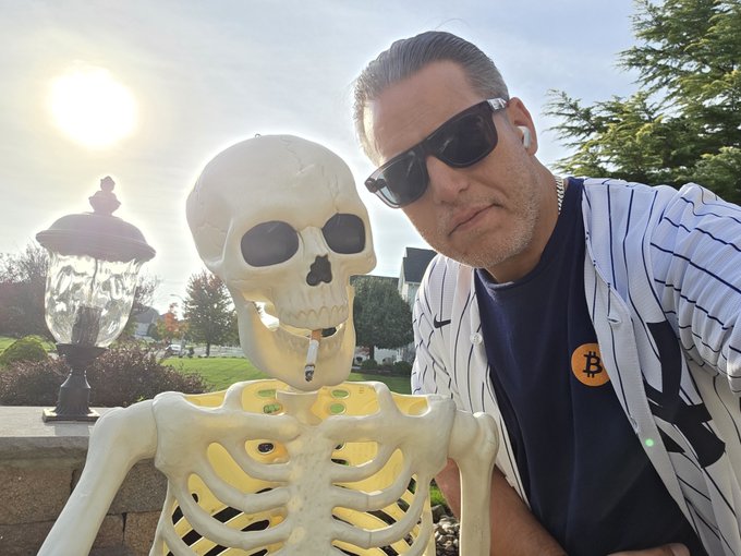 A man wearing sunglasses and a blue shirt with a Bitcoin logo poses next to an illuminated plastic Halloween skeleton prop outdoors.