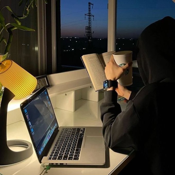 A person wearing a black hoodie sits by a window at dusk, reading a paperback book and holding a Starbucks coffee cup, with a laptop open on the desk beside them.