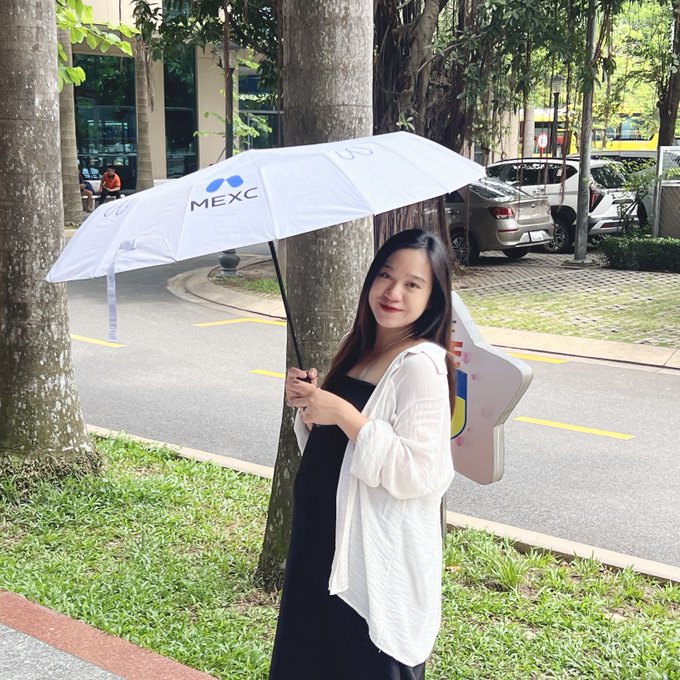 A young woman smiles while holding a white umbrella branded with the "MEXC" logo, seemingly showcasing the promotional item mentioned in the caption. She's dressed in a black outfit with a white cardigan, standing on a tree-lined street on a pleasant weekend day.