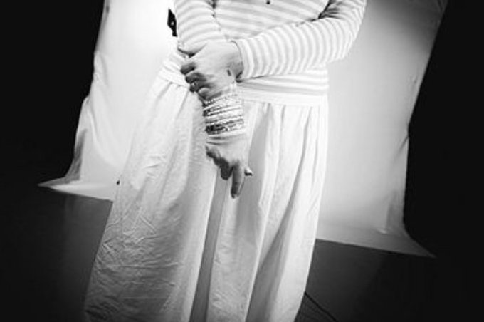 A black and white photograph shows a person from the torso down, wearing a striped shirt and long white skirt, with hands clasped and adorned with multiple bracelets.