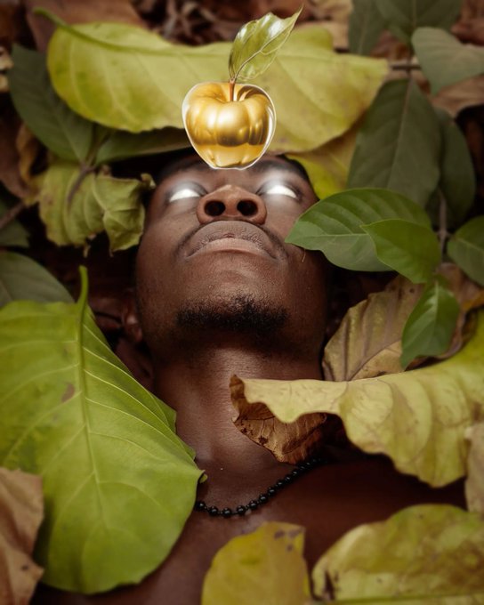 A man lies on his back among green and brown leaves with a golden apple with a leaf balanced on his forehead.