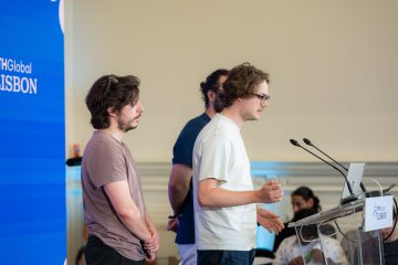 Two young men stand near a microphone and laptop setup in front of a blue banner that partially reads "HGlobal Madison."
