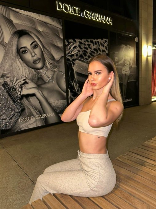 A young woman in a beige crop top and matching pants poses in front of a Dolce & Gabbana storefront featuring large black-and-white advertising posters.