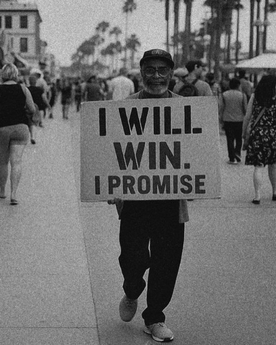 A black-and-white photograph of an older man with a white beard wearing a cap and holding a sign that reads 'I WILL WIN. I PROMISE' while walking down a busy boardwalk lined with palm trees.