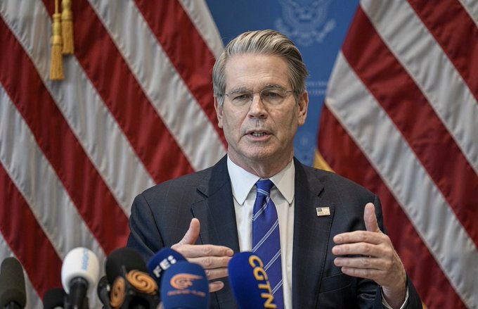 A man in a suit and blue tie speaking at a podium with multiple microphones, standing in front of American flags.