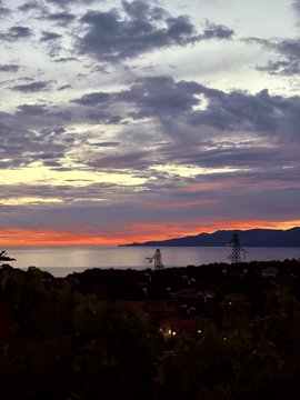 A vertical photo showing a sunset over a calm body of water with silhouetted mountains, power lines, and a coastal town in the foreground beneath a sky filled with purple and orange clouds.