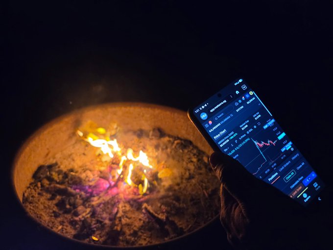 A hand holds a smartphone displaying a cryptocurrency trading app with a 'Zeta Chain' chart next to a glowing fire pit at night.