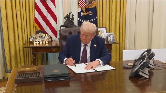 President Donald Trump, wearing a dark suit and purple tie, signs an official document at the Resolute Desk in the Oval Office, with the American flag and presidential seal visible behind him, illustrating his post announcing that "AMERICA WILL LEAD ON CRYPTO GLOBALLY" as he puts pen to paper on what appears to be a related executive action.