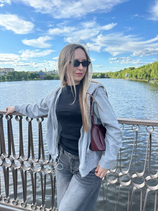 A photograph of a blonde woman wearing sunglasses, a black top, an open gray hoodie, and jeans, standing beside a decorative railing by a river.
