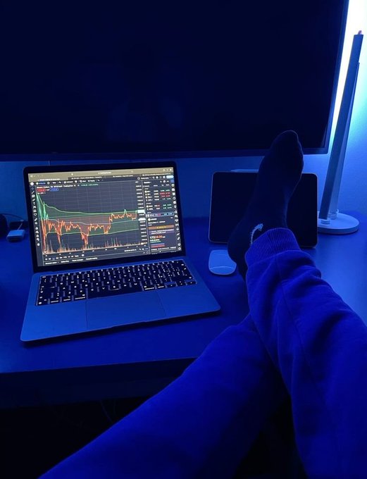 A person relaxes with their feet up on a desk in blue ambient lighting, viewing a stock trading chart on a MacBook displaying green and red candlestick patterns.