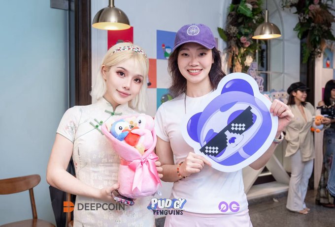 Two women pose together holding a Pudgy Penguins logo cutout and a plush penguin toy, with watermarks for 'DEEPCOIN' and 'PUDGY PENGUINS' visible at the bottom.