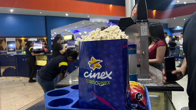 Large blue popcorn bag labeled 'cinex TUDERSCINEX' filled with popcorn on a movie theater counter, with Pepsi cups and people in the background.