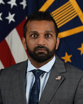 A man with a beard, wearing a gray suit with an American flag pin and patterned tie, stands in front of the U.S. flag and what appears to be the FBI flag. The image accompanies a report that FBI Director Kash Patel disclosed holdings of up to $250,000 in Strategy (ticker: $MSTR).