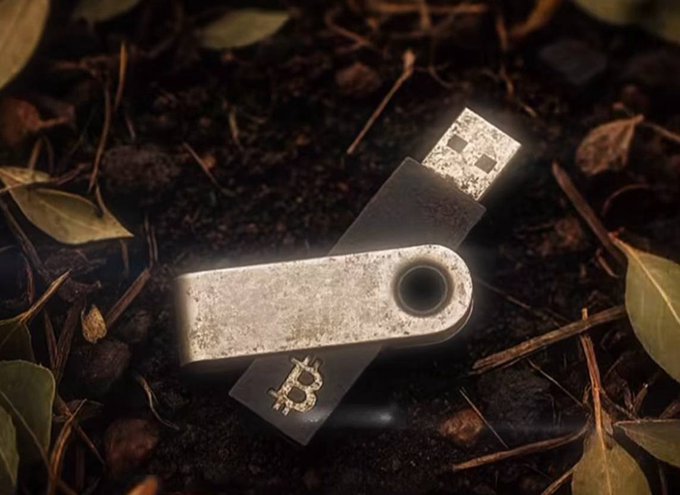 A weathered USB drive and a metal tag with a Bitcoin logo lie on dirt and leaves, with a glowing light effect cast on the device.