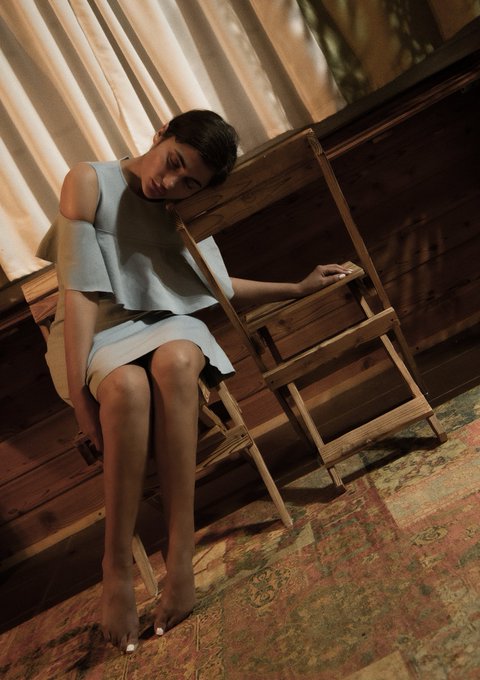 A woman in a light blue dress sits barefoot on a wooden chair, her head tilted to one side, in a softly lit room with curtains and a patterned rug.