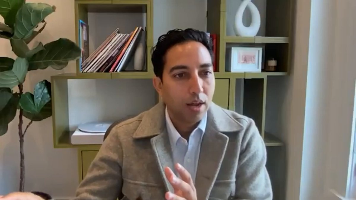 The image shows an adult male sitting at a desk with his hands up, possibly gesturing or making a point during a conversation. The man appears to be wearing a gray jacket and has a potted plant next to him. In front of the man is a bookshelf filled with various items such as books and vases. There are also several books on the shelf, indicating that he might have an interest in reading or studying.