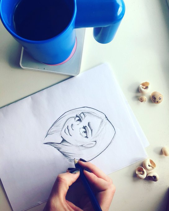 A hand sketches a portrait of a person with short hair on white paper using a pencil, with a blue ceramic mug and scattered cashew nuts resting on the table nearby.