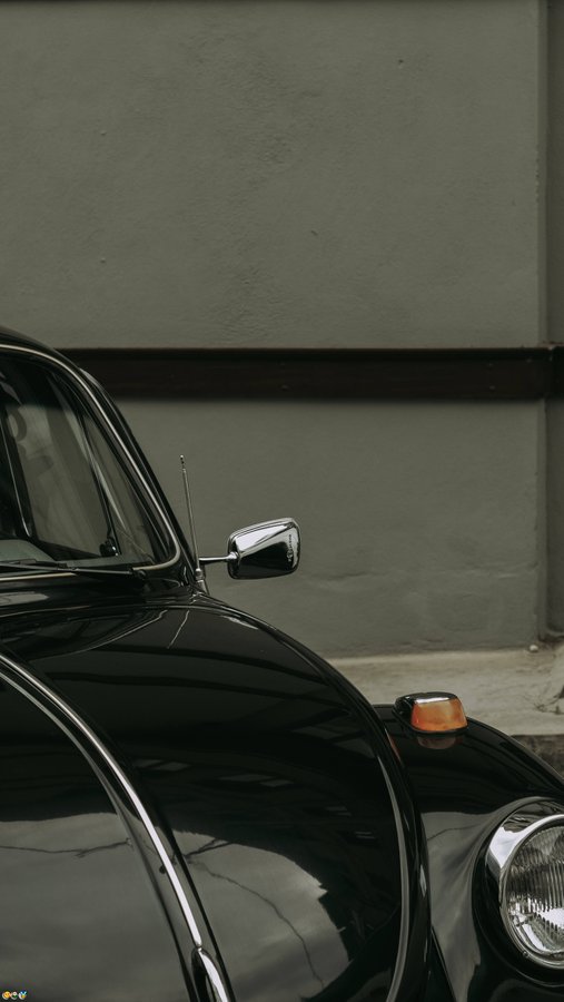 A glossy black vintage Volkswagen Beetle is parked against a gray wall, with its side mirror, antenna, and headlight prominently visible in the shot.