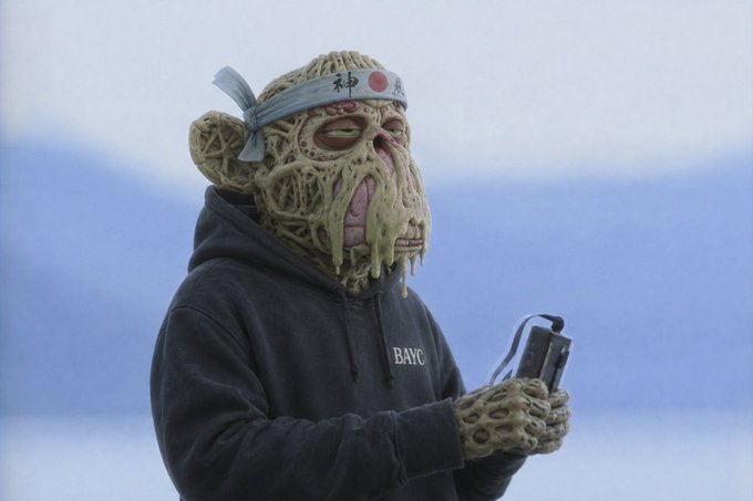 A figure wears a grotesque, web-like green mutant ape mask and matching gloves with a headband featuring the Japanese flag and "神" (god) character, paired with a black hoodie reading "BAYC." Holding a video camera against a hazy blue backdrop, the image embodies the post's celebration of Bored Ape Yacht Club (BAYC) membership and the unique mutated character identity central to the NFT community.