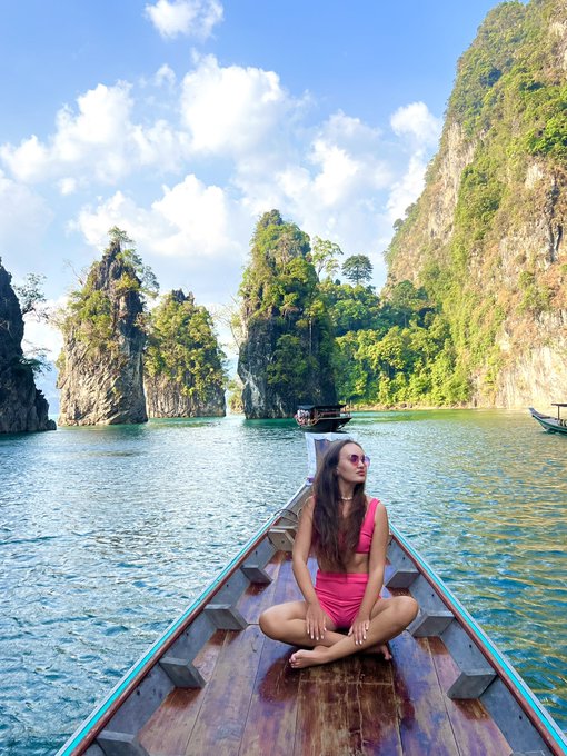 A woman in a pink bikini sits cross-legged at the bow of a wooden boat, floating on turquoise water surrounded by dramatic limestone karst cliffs.