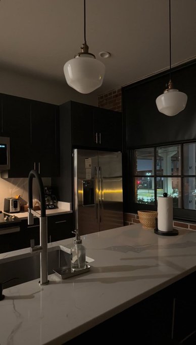 The image shows a kitchen with white countertops and black cabinets. There are two lights hanging above the sink area, providing illumination for various tasks such as washing dishes or preparing food. The kitchen also features a refrigerator, microwave, oven, and other appliances that contribute to its functionality. A window is present in the room, allowing natural light to enter during daytime hours.