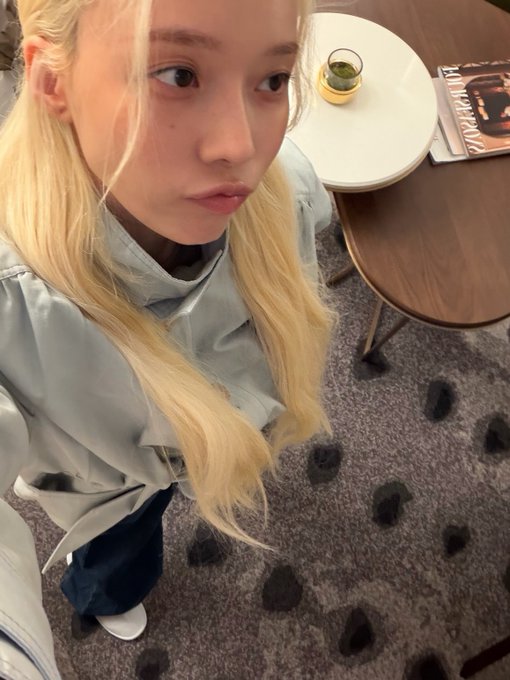 A selfie of a person with long blonde hair wearing a light blue jacket, standing in a room with a wooden table that has a small plant and a magazine on it, on a carpet with dark spots.