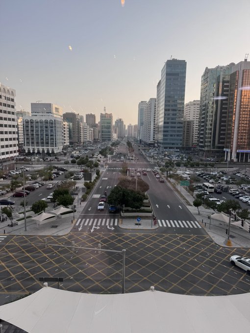 The image shows an elevated view of a wide, modern boulevard in Abu Dhabi with multiple lanes, crosswalks, and a yellow-hatched intersection, flanked by tall glass skyscrapers and office buildings under a hazy sky. This urban cityscape captures the contemporary skyline setting for an upcoming interview with CZ, highlighting Abu Dhabi's distinct modern architecture that contrasts with Dubai.