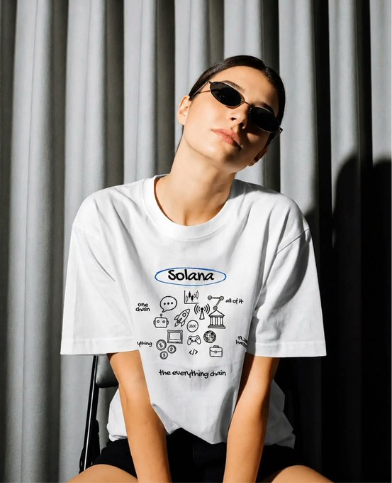 In the image, there's a woman wearing a white t-shirt with a drawing of different objects such as coins and a building. The woman is seated or kneeling down and appears to be posing for a picture. A pair of sunglasses can also be seen on her head. The overall setting seems casual, possibly indoors at home or in an office environment. The image's caption highlights the ease of obtaining this t-shirt through ordering it online from Solana Summit.