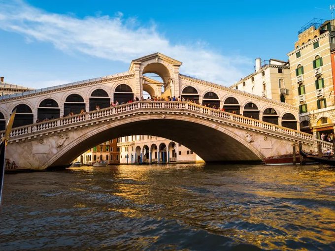 The Rialto Bridge in Venice, Italy, is shown spanning the Grand Canal with its iconic arched stone design, shops, and crowds of tourists under a partly cloudy blue sky.