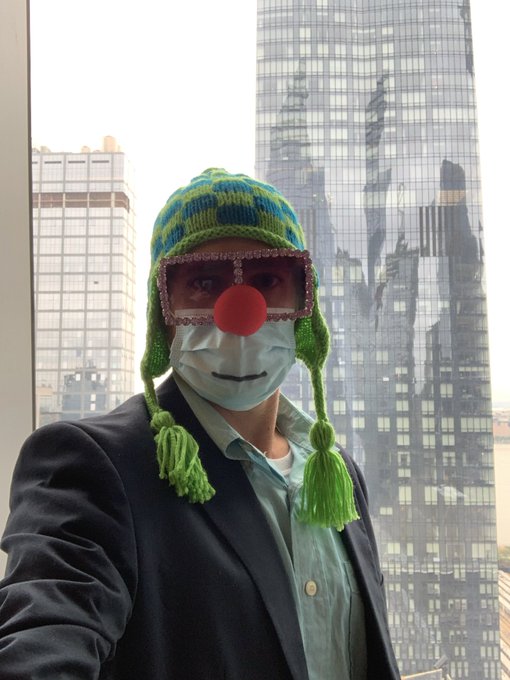 A person wearing a business suit and face mask poses for a selfie with a city skyline in the background, accessorized with a humorous green knit hat featuring tassels, rhinestone-rimmed sunglasses, and a red clown nose.
