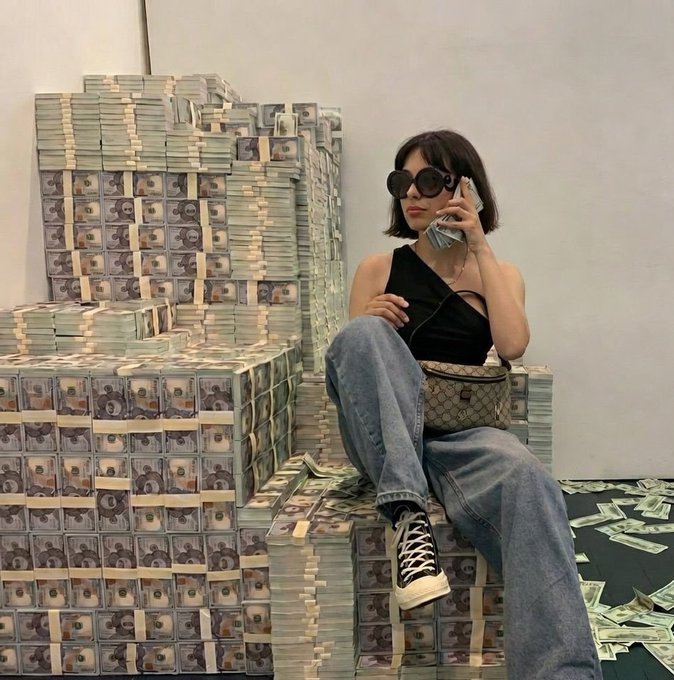 A woman in sunglasses, a black top, and baggy jeans poses on stacks of banded cash bundles while talking on her phone, with a Gucci handbag beside her—visually celebrating her 20 Tezos earnings in classic "money throne" fashion.