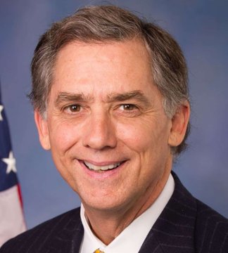 An official portrait of a smiling older man in a dark suit and yellow tie, with an American flag visible in the background.