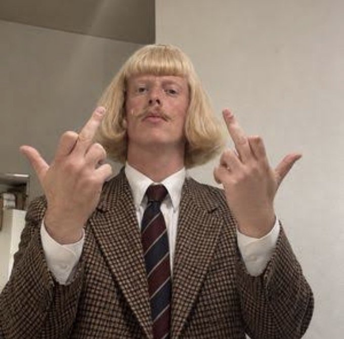 A man wearing a blonde bobbed wig, a mustache, and a brown tweed suit with a striped tie, gesturing with both middle fingers raised.