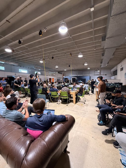 A crowded indoor event space with people seated at round tables and on chairs, watching a presentation or gathering, with some individuals taking photos.