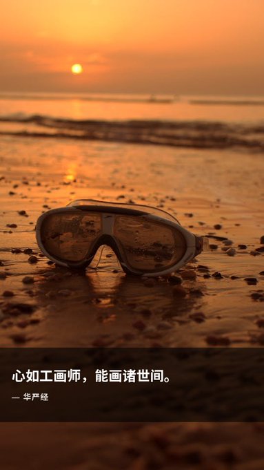 A pair of ski goggles rests on wet sand at a beach during a golden orange sunset, with Chinese text overlay reading '心如工画师，能画诸世间。— 华严经' (The mind is like an artist who can paint all worlds - Flower Garland Sutra).