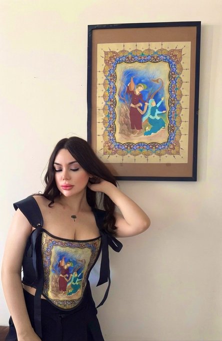 A young woman poses in front of a framed Persian/Rumi-style illuminated manuscript artwork, wearing a corset top that is printed with the same artwork design as the painting behind her.