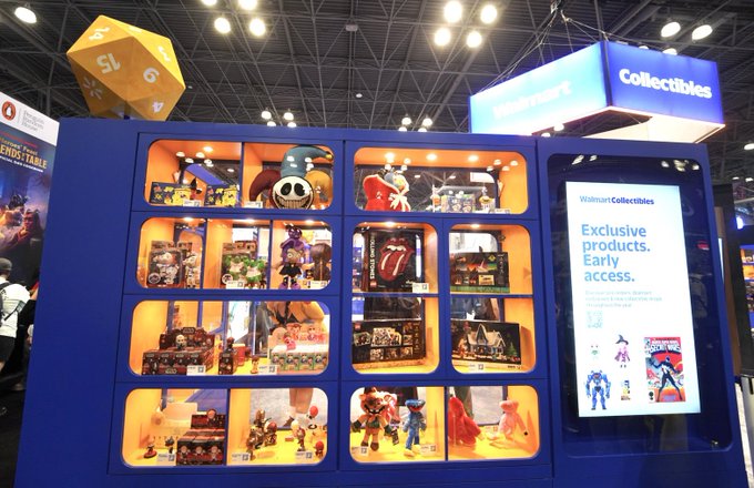 A retail display at a Walmart 'Collectibles' booth showcases various merchandise including figurines, apparel, and a large die prop, with signage advertising 'Walmart Collectibles Exclusive products. Early access.'