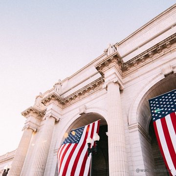 A low-angle photograph of a grand classical building with large arched entrances, featuring two American flags hanging from the arches.