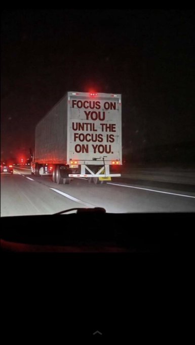 A nighttime highway view of a truck ahead displaying the motivational text 'FOCUS ON YOU UNTIL THE FOCUS IS ON YOU' on its rear doors.