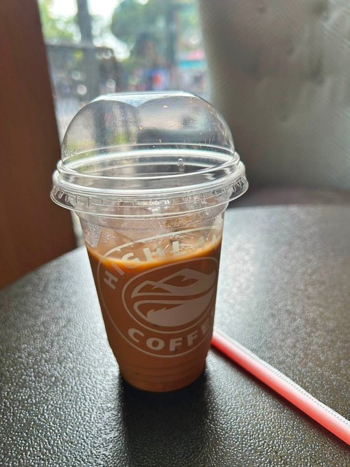 A takeout cup of iced coffee from Highlands Coffee, labeled with the brand's logo, sits on a table with a pink straw beside it. The post describes the poster's afternoon café break during a market trip and their dilemma about reaching 500k to redeem a Pay program reward.