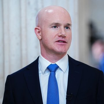 A bald man wearing a dark suit, white shirt, and bright blue tie is shown in a close-up, appearing to speak or be interviewed.