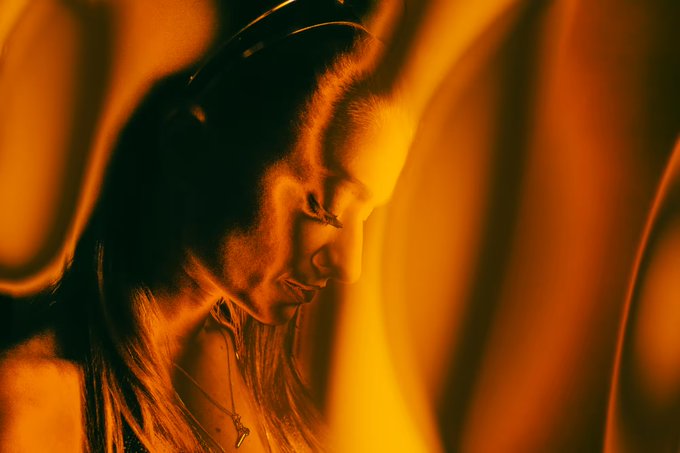 A side profile portrait of a woman bathed in warm orange tones, her gaze directed downward against a softly blurred background, complementing the post's fiery, high-energy "gm" greeting and taco celebration.