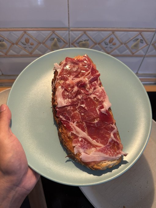 A hand holding a light blue plate with a slice of bread topped with thinly sliced cured meat, likely jamón ibérico, against a kitchen tile backsplash.