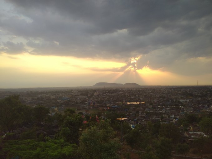 Dramatic crepuscular rays break through dark clouds over a sprawling cityscape, illuminating distant twin hills on the horizon during a golden sunset. The elevated view captures lush green foreground trees giving way to an expansive urban landscape bathed in warm evening light, perfectly matching the peaceful weekend mood and "touching mountain" sentiment of the post.
