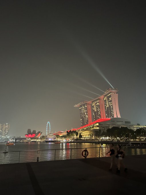 A nighttime cityscape features the illuminated Marina Bay Sands hotel in Singapore with its light beams shooting into the sky, and a Ferris wheel visible across the water.