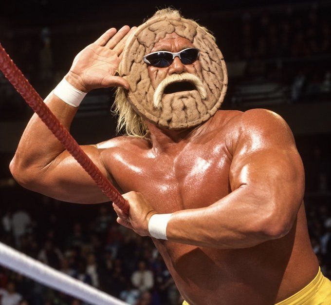 A muscular blonde wrestler in yellow trunks poses in a wrestling ring, holding a rope while cupping his ear, wearing his signature brain-shaped mask.