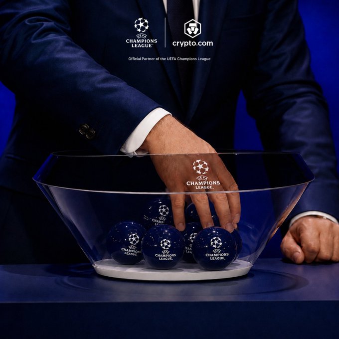 A suited individual reaches into a glass bowl containing blue "UEFA Champions League" branded balls during a tournament draw ceremony, with the Crypto.com logo displayed as the official partner of the UEFA Champions League at the top. The image captures the anticipation and excitement of the new Champions League season draw referenced in the post.